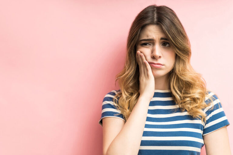Person with long wavy hair in a blue and white striped shirt holding their cheek in discomfort, standing against a pink background—suggesting facial pain possibly related to a root canal.