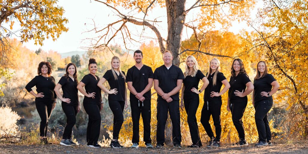 A group of ten individuals dressed in black uniforms stands outdoors in front of autumn-colored trees. The setting features yellow and orange foliage, suggesting a fall season.