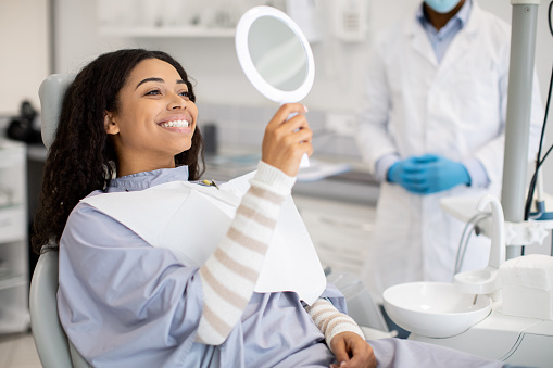 Patient seated in a dental chair wearing a bib, holding a mirror to check their teeth, with a dentist in gloves standing nearby in a clean clinic setting.