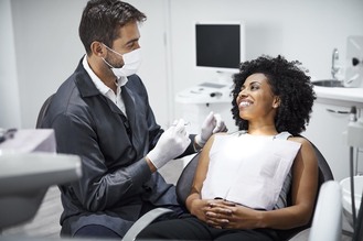 A dental professional in a mask and gloves prepares instruments while a patient reclines in a chair during a consultation in a clinical office.